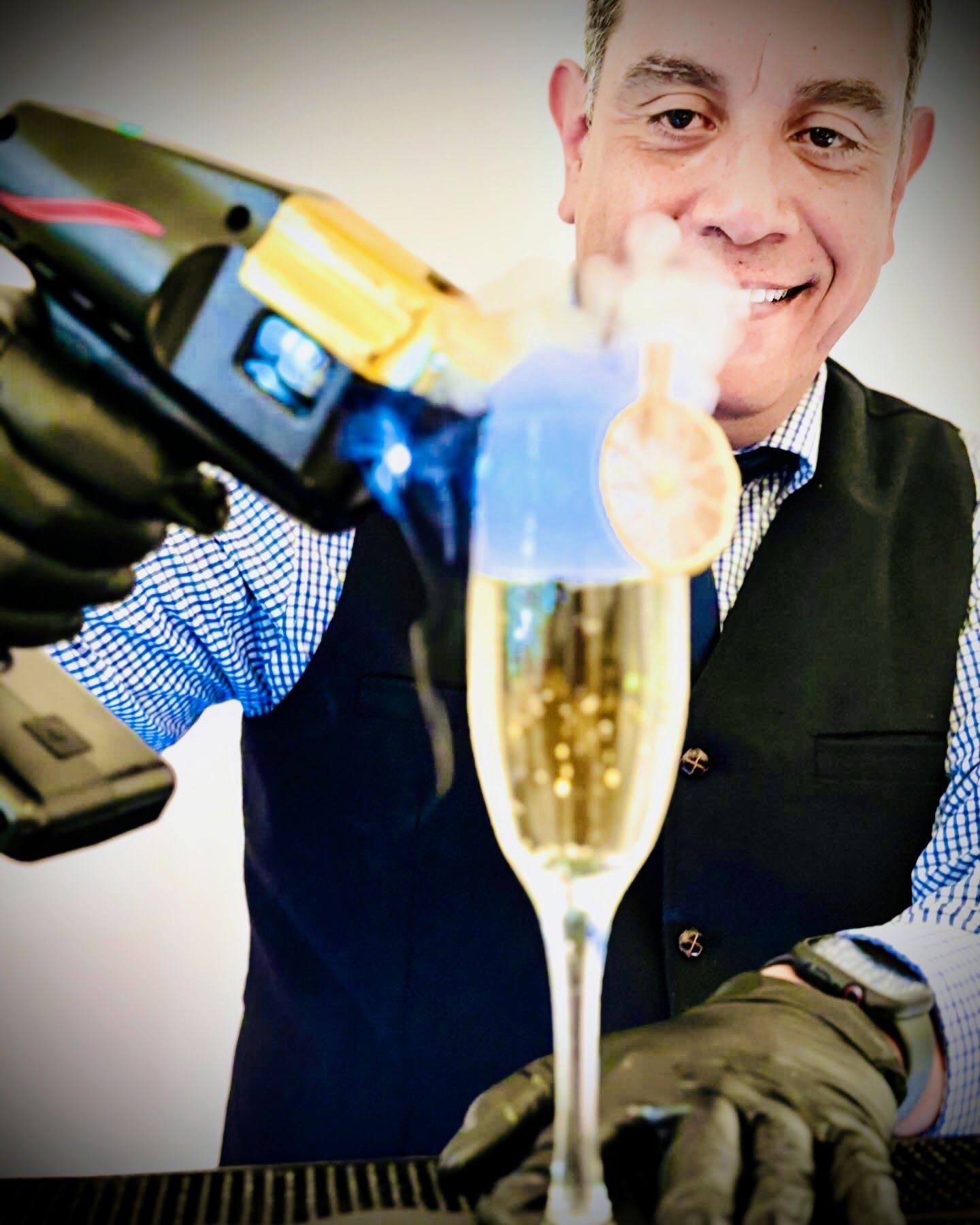 Bartender using a torch to smoke a cocktail, with a smiling face in the background.