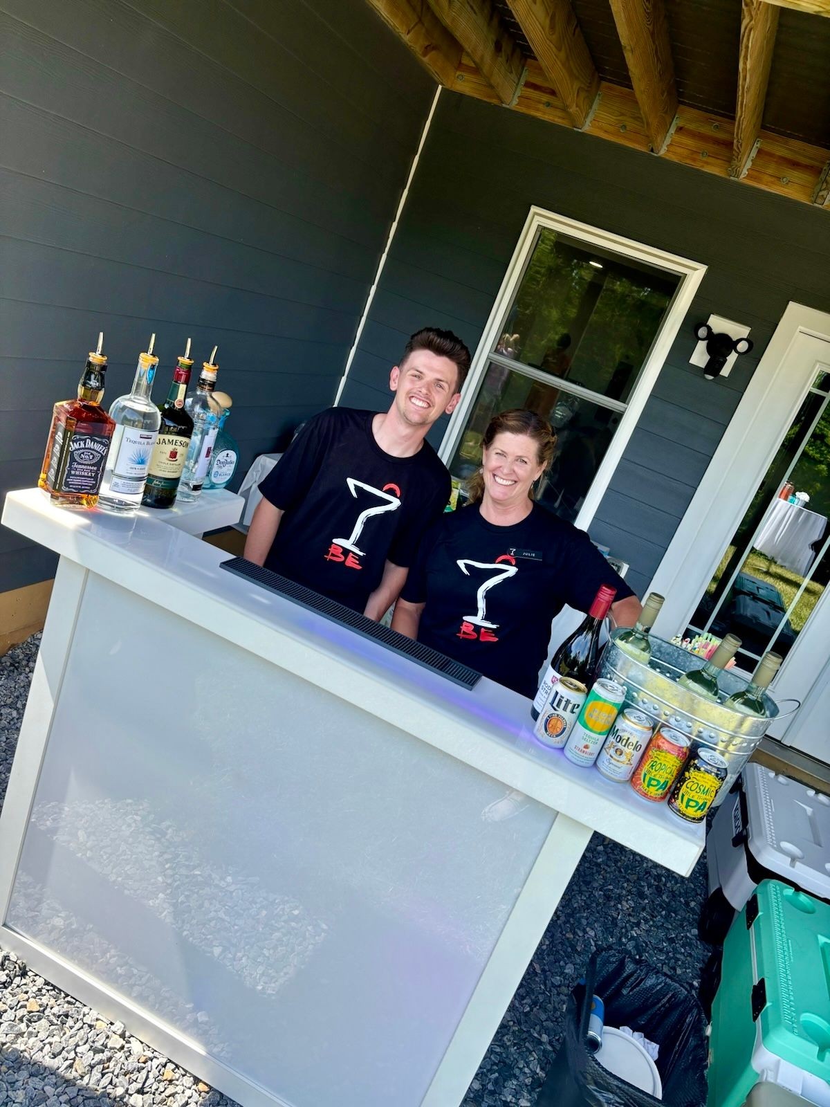 Two people behind a home bar with various liquor bottles and cans on display under a covered patio.